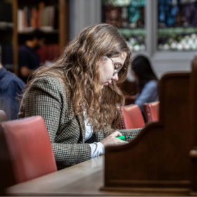 Colour photo of a student writing whilst sat at the desks in the New College Library
